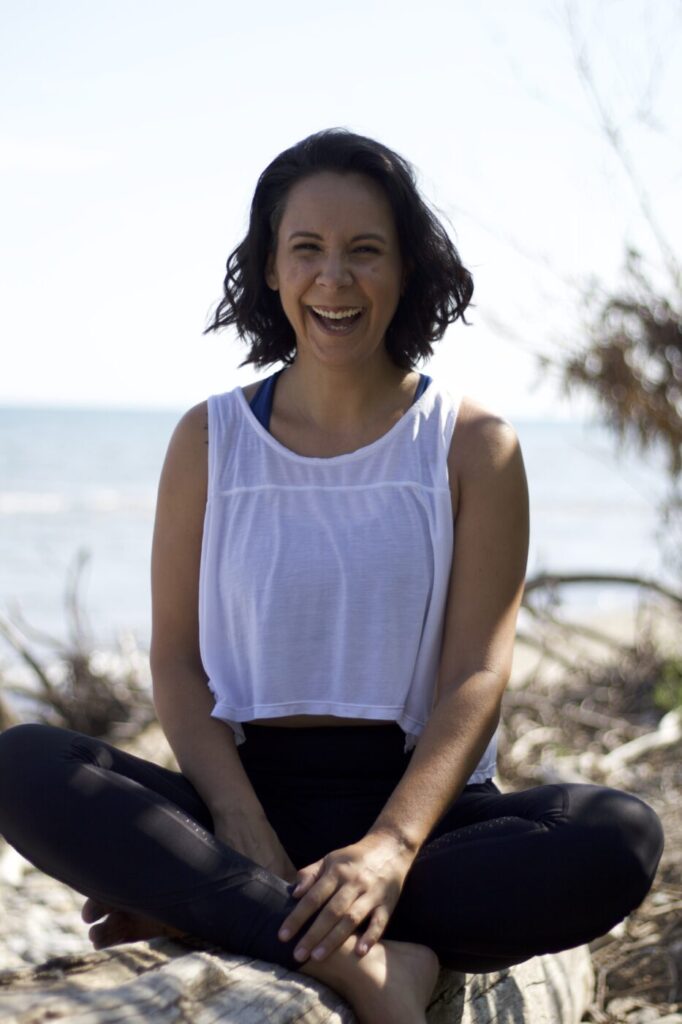 josi-laughing, seated on a log, with water and sky behind them.