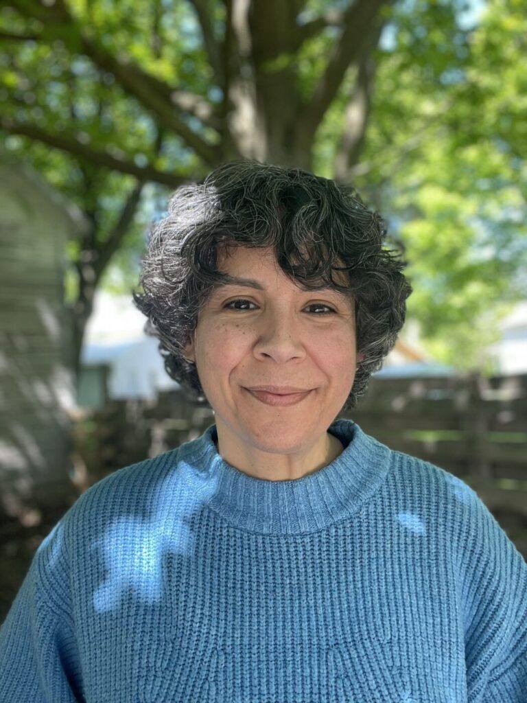 josi stands under a tree, with its trunk behind them, dappled sunlight across their blue sweater. Smiling towards the camera, with a mop of black and grey short curls.